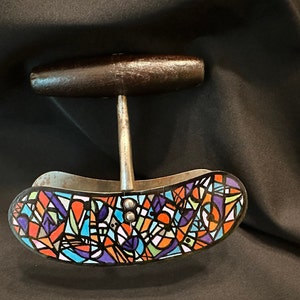 May include: A vintage food chopper with a dark wooden handle and a stainless steel blade. The curved base features a colorful, abstract design with geometric shapes in orange, blue, purple, and green. The chopper is likely used for mincing or chopping food items.