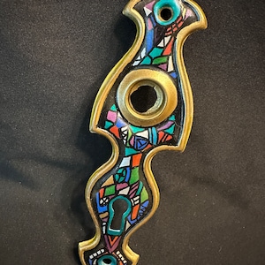 May include: A brass door handle with a black background and colorful geometric design. The handle has a keyhole and two round holes.