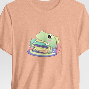 May include: Peach-colored t-shirt featuring a cartoon frog enjoying a burger, popcorn, and a drink. The frog is sitting on a colorful rug with pillows. The design is playful and whimsical, perfect for casual wear.