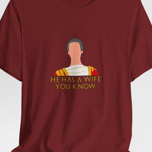 May include: Burgundy t-shirt featuring a cartoon illustration of a figure in Roman attire. The text "HE HAS A WIFE YOU KNOW..." is printed in gold and white. The shirt is made of a soft material.