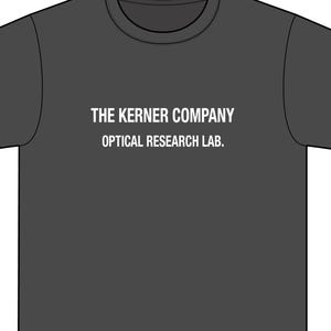 May include: Dark gray t-shirt with the text "THE KERNER COMPANY OPTICAL RESEARCH LAB." printed in white. The shirt has a crew neck and short sleeves. The text is centered on the front of the shirt.