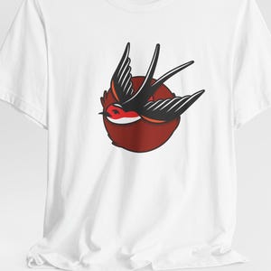 May include: White t-shirt featuring a stylized tattoo-style swallow design. The swallow is black with red accents, set against a red circular background. The design is centered on the front of the shirt.