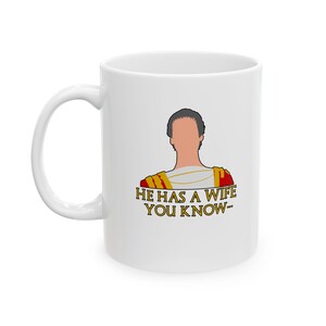 May include: A white ceramic mug featuring a graphic illustration of a person wearing a red and gold Roman-style tunic. Below the illustration, the text reads, "HE HAS A WIFE YOU KNOW..." in gold lettering with a dark outline on the front surface.