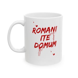 May include: A white ceramic mug featuring the red text ROMANI ITE DOMUM printed in a distressed, brush-stroke style. The lettering appears to have a splattered, paint-like effect with small red droplets surrounding the words on the mug surface.