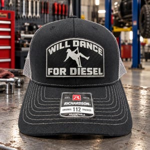 May include: A black trucker hat with a white mesh back and a rectangular patch on the front. The patch features a silhouette of a person dancing on a pole with the text WILL DANCE FOR DIESEL. A sticker on the brim reads RICHARDSON ORIGINAL 112 TRUCKER.