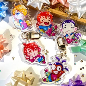 May include: Five colorful cartoon character keychains with gold chains. The characters are a girl with blonde hair, a girl with red hair, a girl with purple hair, a girl with red hair, and a boy with green hair. The keychains are on a white surface with gold stars.
