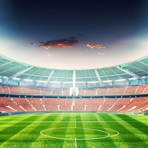 May include: An empty soccer stadium with red seats and a green field. The stadium is lit by bright lights and the sky is blue with a few clouds.
