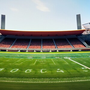 May include: Panoramic view of a football stadium with a green field and white yard lines. The stands are filled with red seats under a red and orange roof. Two tall, dark pillars stand at the stadium's edge.