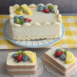 May include: A rectangular white cake with decorative frosting, topped with lemon slices, blueberries, and strawberries, sits on a blue plate. Two smaller square cakes with similar decorations rest on doilies. The cakes are on a yellow and white checkered surface.