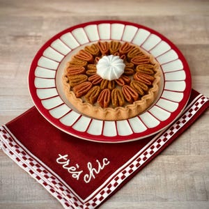 May include: A pecan pie with a dollop of whipped cream sits on a red and white plate. The plate rests on a red napkin embroidered with "très chic" in white. The pie crust is golden brown, and the pecans are arranged in a circular pattern.