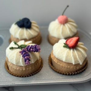 May include: Four felt tarts with white frosting and various toppings, including blueberries, a cherry, a strawberry, and lavender. The tarts sit in a gray baking pan, showcasing the handmade details and vibrant colors.