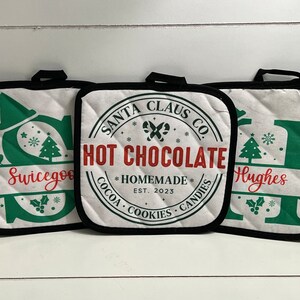 May include: Three white quilted oven gloves with black trim. The centre oven glove has a red and black circle with the text "SANTA CLAUS CO. HOT CHOCOLATE *HOMEMADE* EST. 2023 COCOA COOKIES CANDIES*" The left oven glove has a green letter "S" with the text "Swicegoo" and the right oven glove has a green letter "I" with the text "Hughes".