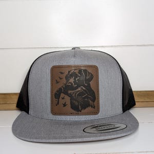 May include: A gray and black trucker hat with a brown leather patch featuring a dog holding a duck. The hat has a mesh back and a curved brim. The patch has a detailed illustration of the dog and ducks.