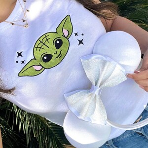 Op de afbeelding: Wit T-shirt met een groen cartoongezicht en zwarte ogen. Het shirt wordt gecombineerd met witte muizenoren met een sprankelende strik. De oren worden vastgehouden door een persoon die het shirt draagt.