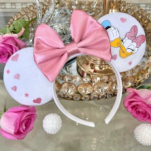 May include: White headband with pink satin bow and round white ears. One ear features a cartoon image of Daisy Duck and Donald Duck. The other ear has pink glitter hearts and stars. Pink roses and decorative balls surround the headband.