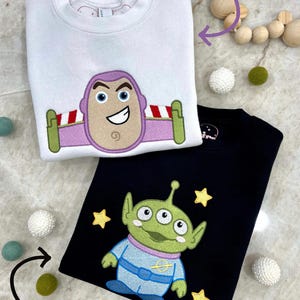K&ouml;nnte beinhalten: Zwei Sweatshirts mit Rundhalsausschnitt, eines wei&szlig; und eines schwarz. Das wei&szlig;e Sweatshirt zeigt ein Buzz Lightyear-Design, w&auml;hrend das schwarze einen gr&uuml;nen Au&szlig;erirdischen mit gelben Sternen zeigt. Die Sweatshirts sind Teil der Kollektion "Space Traveler" und "Space Friend".