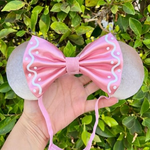 May include: A pink satin bow headband with gray ears. The bow features white wavy lines and dots. The headband is held up by a pink elastic band. The background is green foliage.