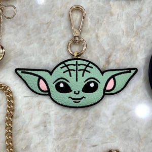 May include: A light green keychain featuring a cartoon character's face with large ears, black eyes, and pink accents. The keychain has a gold-colored clasp and ring. The character's face is stitched onto a black border.