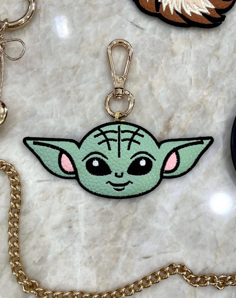 May include: A light green keychain featuring a cartoon character's face with large ears, black eyes, and pink accents. The keychain has a gold-colored clasp and ring. The character's face is stitched onto a black border.