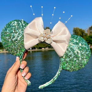 Może przedstawiać: Para uszu myszki z zielonymi cekinami, z dużą kremową kokardą i kwiatową ozdobą pośrodku. Opaska jest miętowo-zielona i ozdobiona cekinami. Delikatne perłowe akcenty rozciągają się od kokardy.