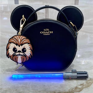 May include: A navy blue Coach purse with a gold zipper and handle, featuring a charm of a cartoon character and a blue lightsaber. The purse has two circular ear-shaped accents. The charm is brown and tan. The text at the bottom says "Bag Charm only - Purse is not included".