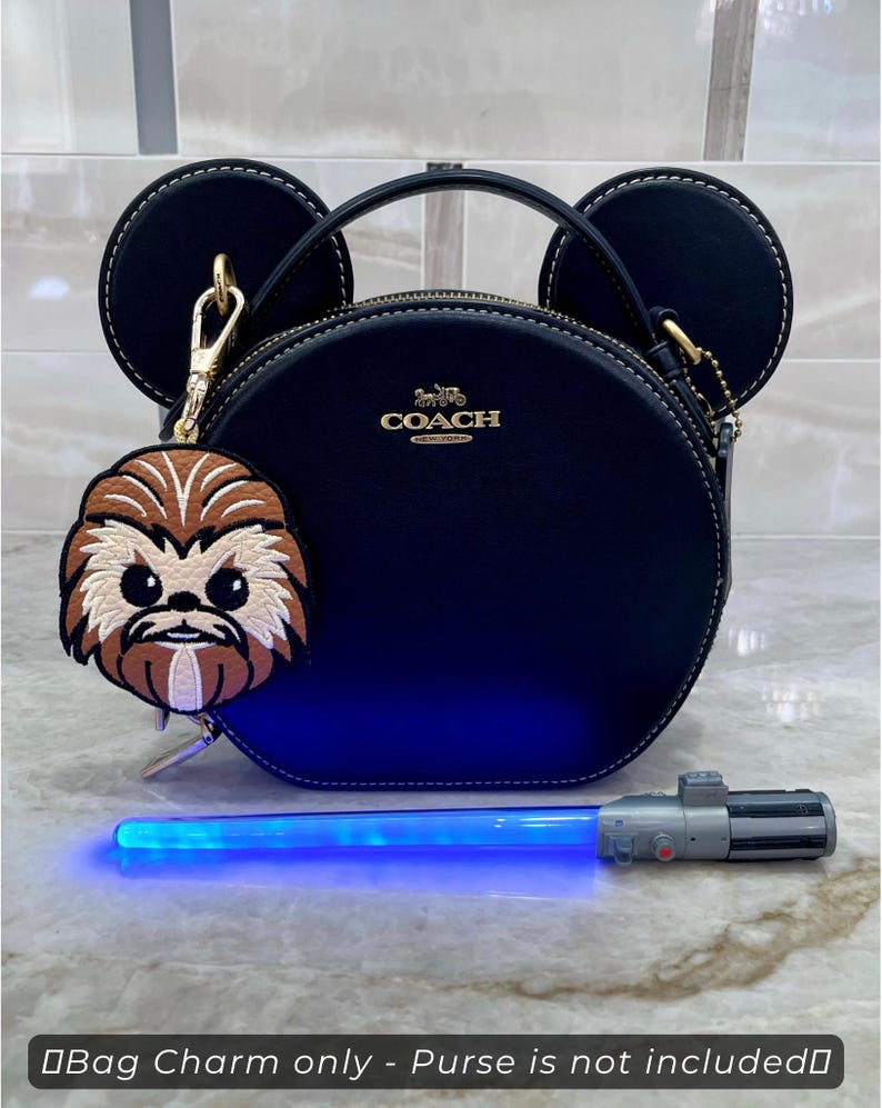 May include: A navy blue Coach purse with a gold zipper and handle, featuring a charm of a cartoon character and a blue lightsaber. The purse has two circular ear-shaped accents. The charm is brown and tan. The text at the bottom says "Bag Charm only - Purse is not included".