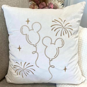 May include: A white square pillow with embroidered gold-colored Mickey Mouse balloon designs and fireworks. The pillow also features gold star accents, creating a festive and celebratory theme. The pillow is made of a soft, plush material.
