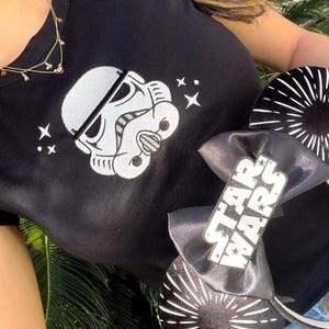 May include: Black t-shirt featuring a white Stormtrooper helmet design and star accents. A black bow with "Star Wars" text and black and white circular ear accessories are also visible.