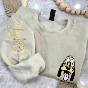 Puede incluir: Una sudadera beige con un "Pluto" bordado en amarillo en la manga y una imagen de dibujos animados de Pluto en la parte delantera.