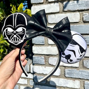 May include: Black and white Star Wars themed Minnie Mouse ears with a black bow. The ears feature Darth Vader on one side and a Stormtrooper on the other.