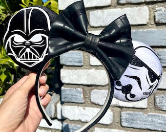 May include: Black and white Star Wars themed Minnie Mouse ears with a black bow. The ears feature Darth Vader on one side and a Stormtrooper on the other.