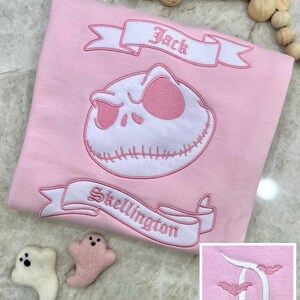 May include: A light pink shirt featuring an embroidered Jack Skellington design with the text "Jack" and "Skellington" on banners. The shirt also has a small embroidered design in the corner. Two small ghost-shaped felt ornaments are next to the shirt.