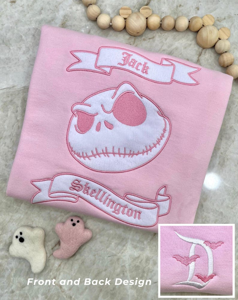 May include: A light pink shirt featuring an embroidered Jack Skellington design with the text "Jack" and "Skellington" on banners. The shirt also has a small embroidered design in the corner. Two small ghost-shaped felt ornaments are next to the shirt.