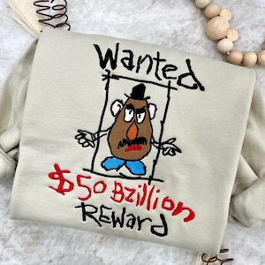 Peut inclure: Un t-shirt beige avec un motif brodé rouge et noir d'une affiche de recherche d'un personnage de tête de pomme de terre de dessin animé. Le texte sur l'affiche indique "Wanted" et "$50 bzillion Reward".