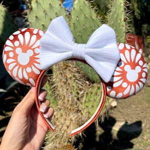 May include: A pair of orange and white mouse ears with a white bow. The ears feature a white daisy pattern.