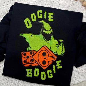 Puede incluir: Camiseta negra con una imagen bordada en verde de Oogie Boogie de Pesadilla antes de Navidad. La imagen incluye dos dados naranjas con diseños de calaveras negras. El texto "Oogie Boogie" está bordado en verde.