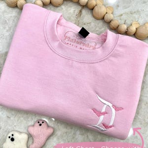 May include: A light pink sweatshirt with an embroidered white and pink letter "D" design and bats. The text "Pixie Pair's Magic Wear" is on the collar. The text "Left Chest - Choose with or without bats!" is on the bottom right.