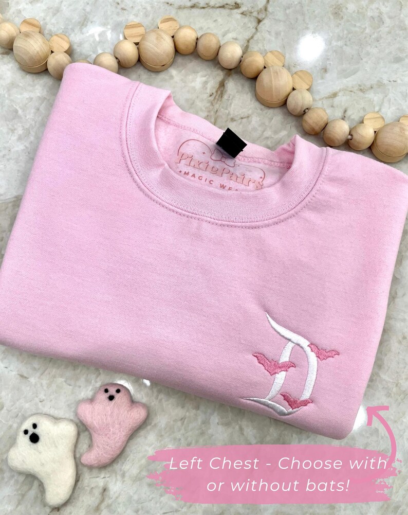 May include: A light pink sweatshirt with an embroidered white and pink letter "D" design and bats. The text "Pixie Pair's Magic Wear" is on the collar. The text "Left Chest - Choose with or without bats!" is on the bottom right.
