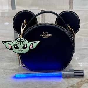 May include: A navy blue Coach purse with black Mickey Mouse ears and a gold Coach logo. A green and pink Baby Yoda bag charm hangs from the purse. A blue lightsaber toy is in front of the purse.