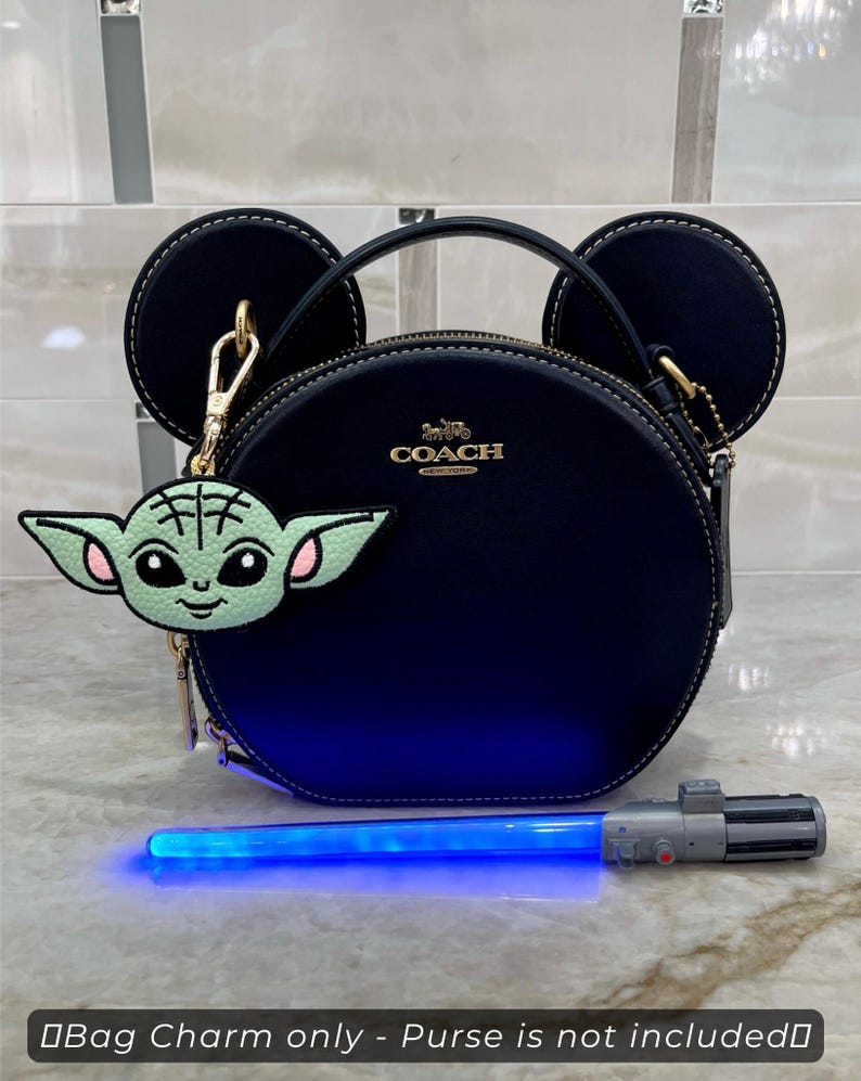 May include: A navy blue Coach purse with black Mickey Mouse ears and a gold Coach logo. A green and pink Baby Yoda bag charm hangs from the purse. A blue lightsaber toy is in front of the purse.