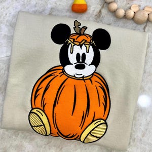 May include: A beige sweatshirt featuring a Mickey Mouse design dressed as a pumpkin. The design includes a black outline, orange pumpkin, and yellow accents. The sweatshirt is decorated with a Halloween theme.