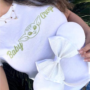 Op de afbeelding: Wit T-shirt met groen geborduurde tekst en een cartoon karakter ontwerp. Een paar witte muizenoren met een sprankelende strik worden voor het shirt gehouden. De tekst op het shirt luidt "Baby Grogu".
