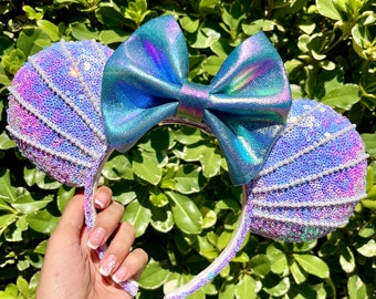 May include: Purple and blue iridescent Minnie Mouse ears with a large iridescent bow. The ears are covered in sequins and pearls.