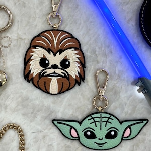 May include: Two character keychains. One keychain features a brown and tan Chewbacca design, and the other features a green and pink Baby Yoda design. Both keychains have gold-colored metal clasps.