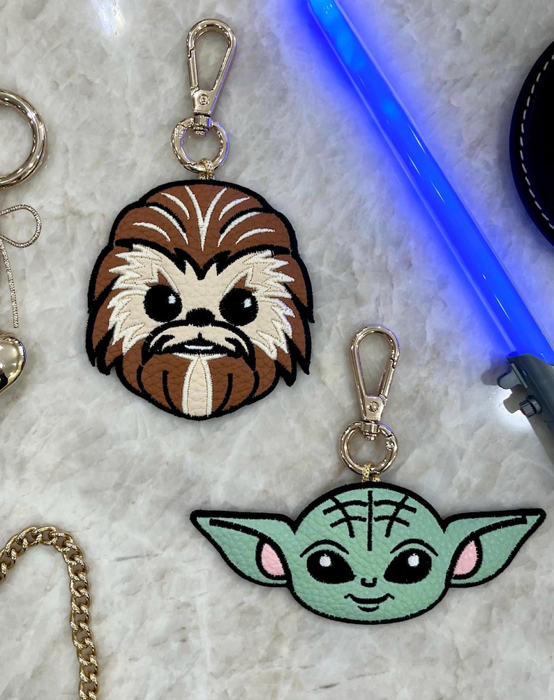 May include: Two character keychains. One keychain features a brown and tan Chewbacca design, and the other features a green and pink Baby Yoda design. Both keychains have gold-colored metal clasps.