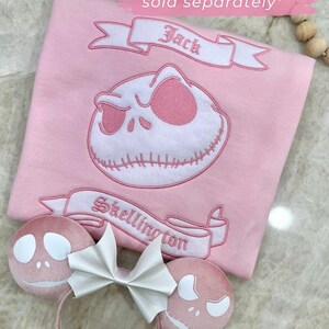 May include: Pink fabric with a Jack Skellington design, banner text "Jack" and "Skellington". Includes "King Jack" ears with a white bow. Text reads "Matching Mouse Ears sold separately".