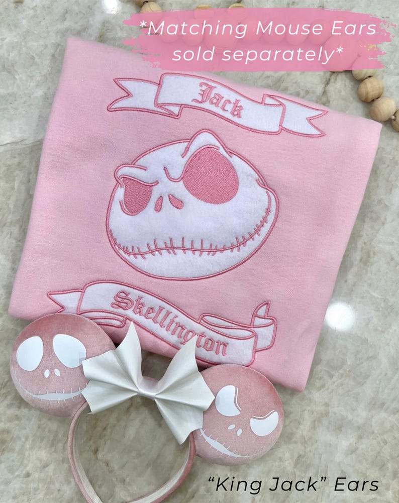 May include: Pink fabric with a Jack Skellington design, banner text "Jack" and "Skellington". Includes "King Jack" ears with a white bow. Text reads "Matching Mouse Ears sold separately".