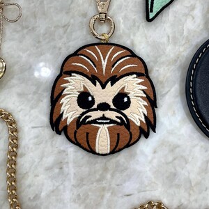 May include: A brown and tan leather keychain featuring an embroidered character face with black eyes and a gold-colored clasp. The keychain has a gold chain and is a fun accessory.