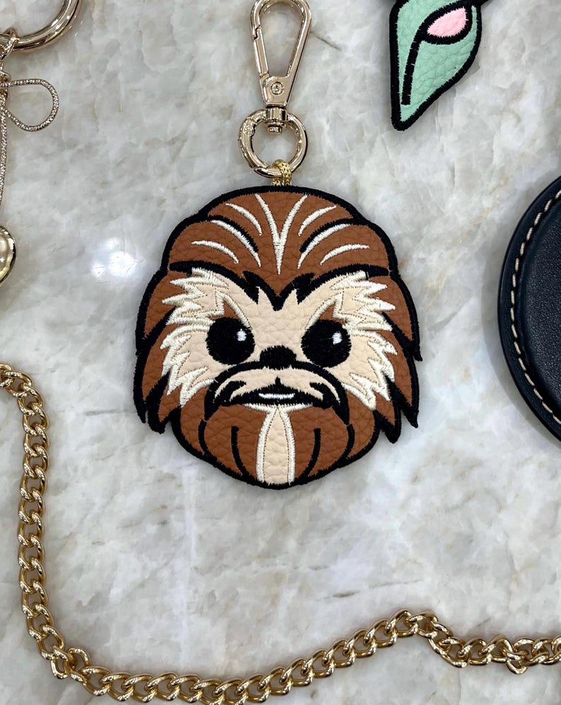 May include: A brown and tan leather keychain featuring an embroidered character face with black eyes and a gold-colored clasp. The keychain has a gold chain and is a fun accessory.