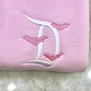 May include: A light pink sweatshirt with a white embroidered letter "D" and three pink bats. The bats are positioned around the letter. The sweatshirt is made of a soft, textured fabric.